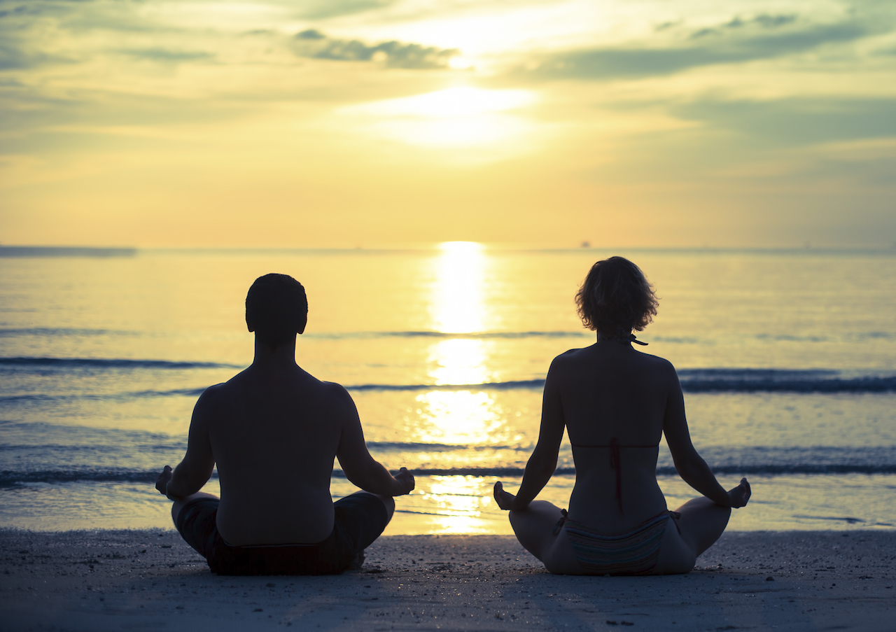 Young couple practicing yoga in the lotus position on the ocean beach during sunset. Cross-process p